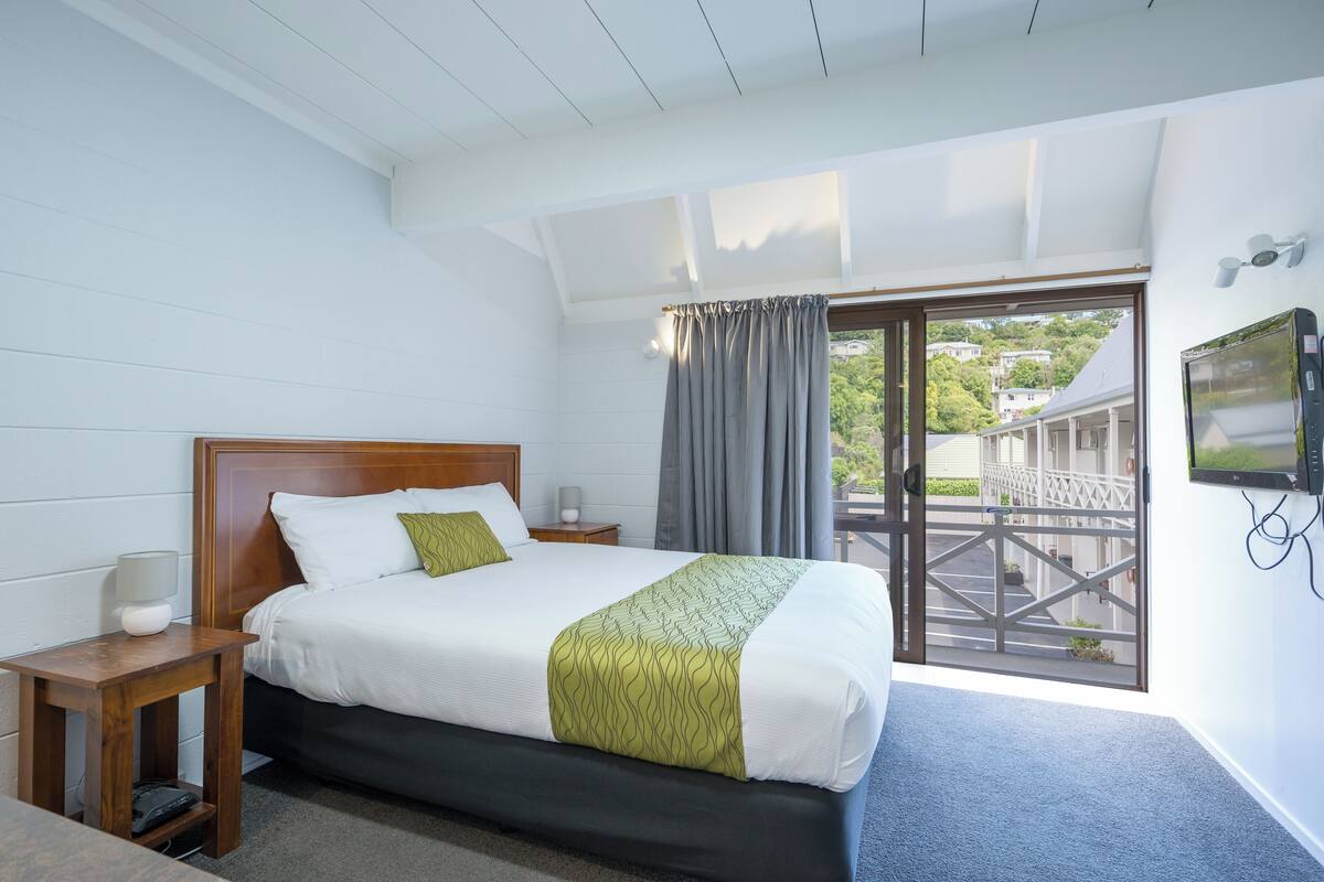 A hotel room with a bed, a nightstand, a TV, and a view of the outside.