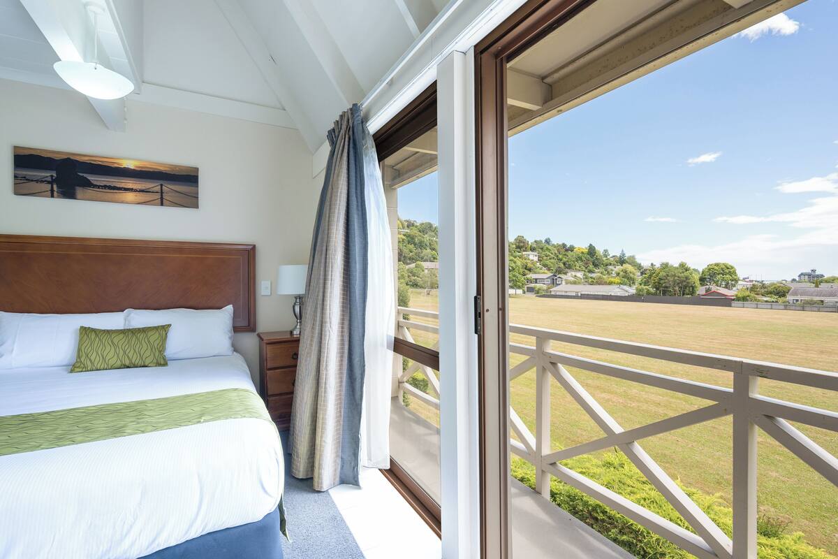 A hotel room with a large bed, a view of a grassy field, and a balcony with a railing.