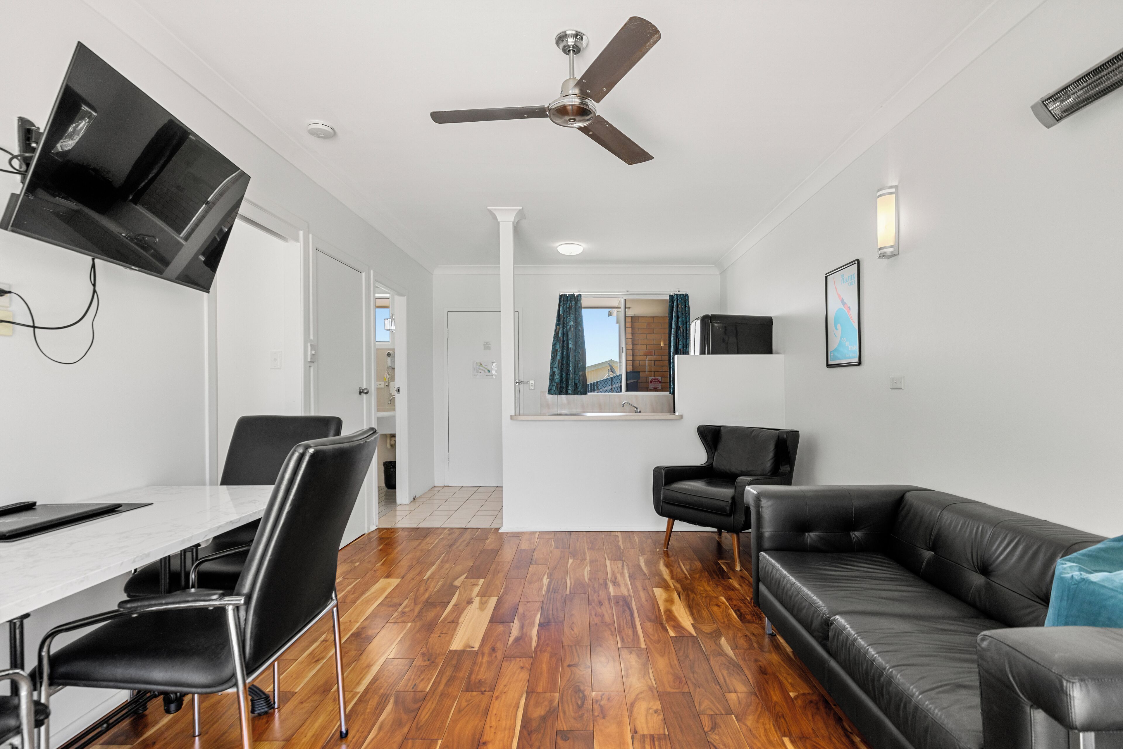 A modern living room with a black leather sofa, a dining table with chairs, a ceiling fan, and a flat-screen TV mounted on the wall.