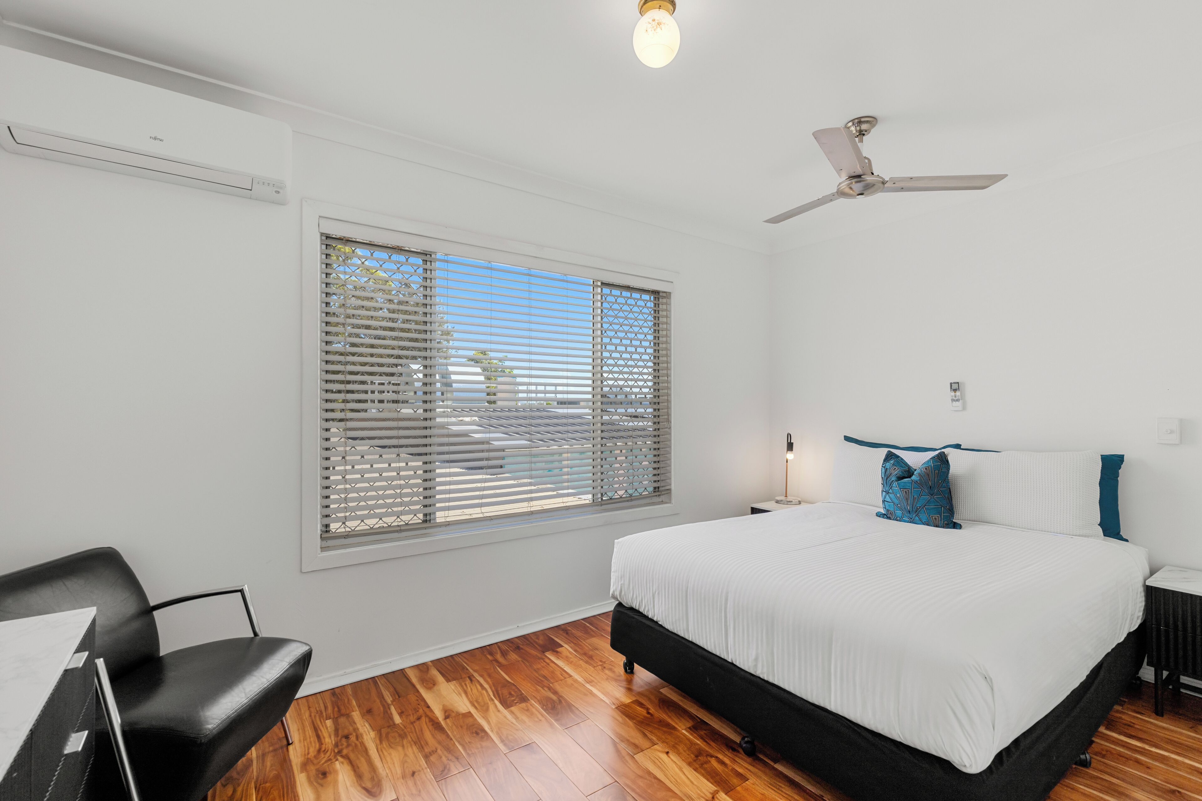 Une chambre à coucher comprenant un lit, un ventilateur de plafond, une fenêtre avec des stores, une chaise et une table.