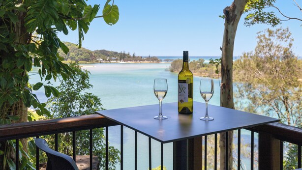 Une table avec une bouteille de vin et deux verres, donnant sur un paysage côtier avec des arbres et un ciel dégagé.