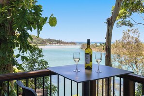 A table with a bottle of wine and two glasses, overlooking a coastal landscape with trees and a clear sky.