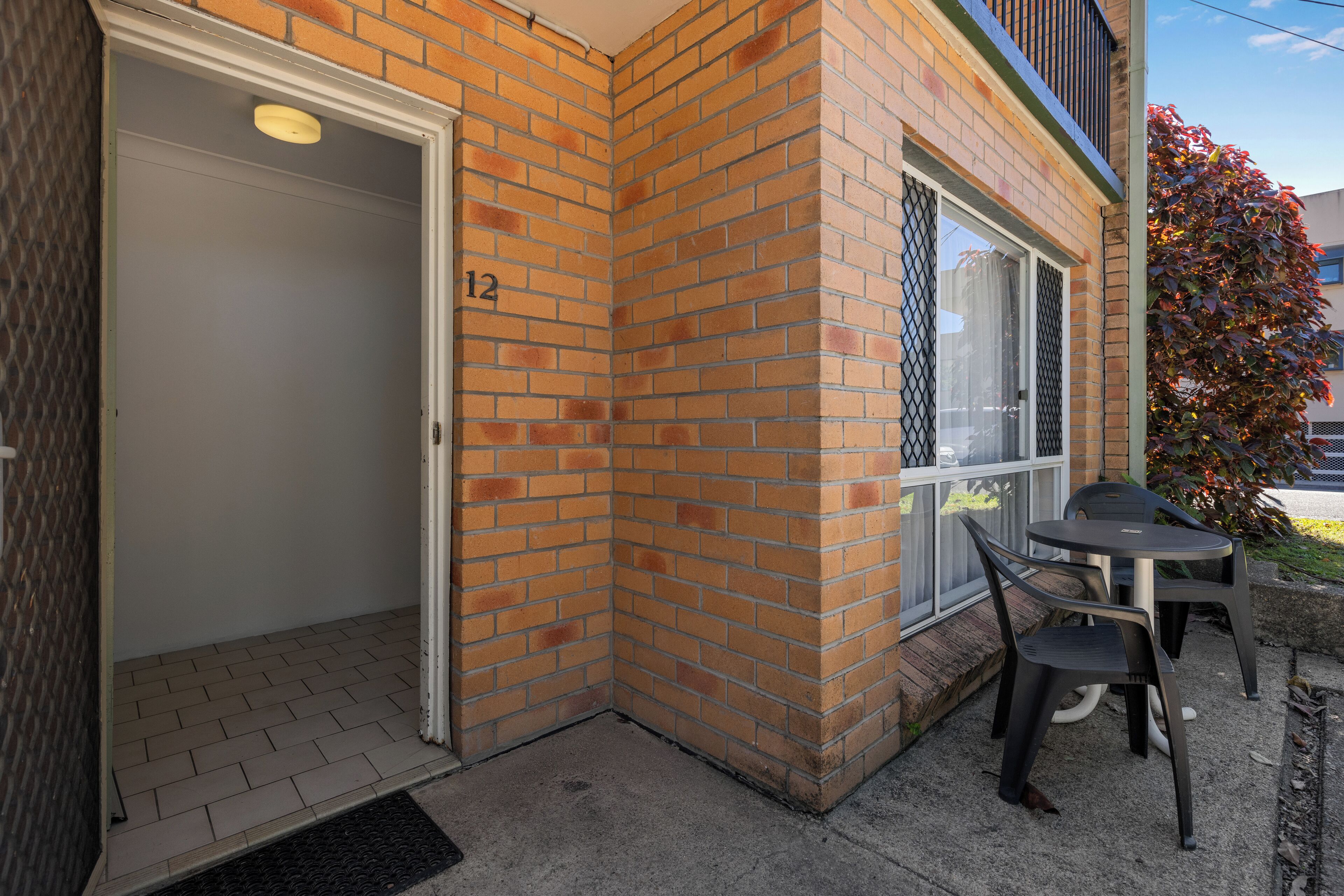 A corner of a building with a brick exterior, a door marked with the number 12, and a small outdoor seating area with two chairs and a table.