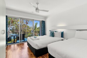 Habitación de hotel con dos camas, ventilador de techo, balcón con mesa y sillas, y vista a palmeras.