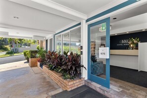 Fachada de hotel en azul y blanco, con un mostrador de recepción y una jardinera con diversas plantas.