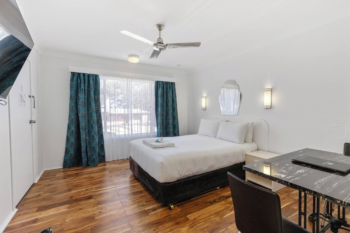 A hotel room with a bed, a desk, a chair, a ceiling fan, and a window with curtains.