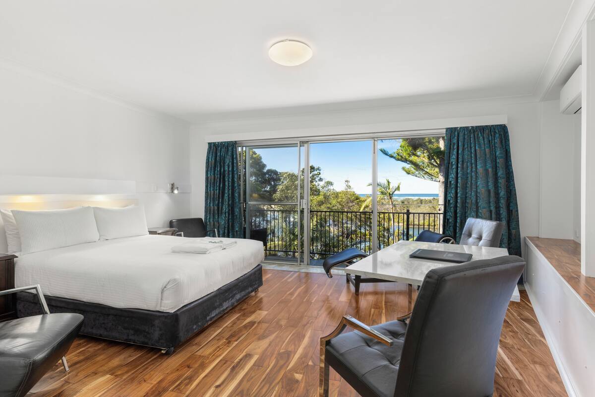 A hotel room with a large bed, a dining table, and a balcony with a view of the sea.
