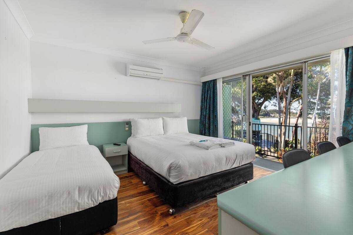 A hotel room with two beds, a ceiling fan, an air conditioning unit, a desk, and a view of the outdoors.