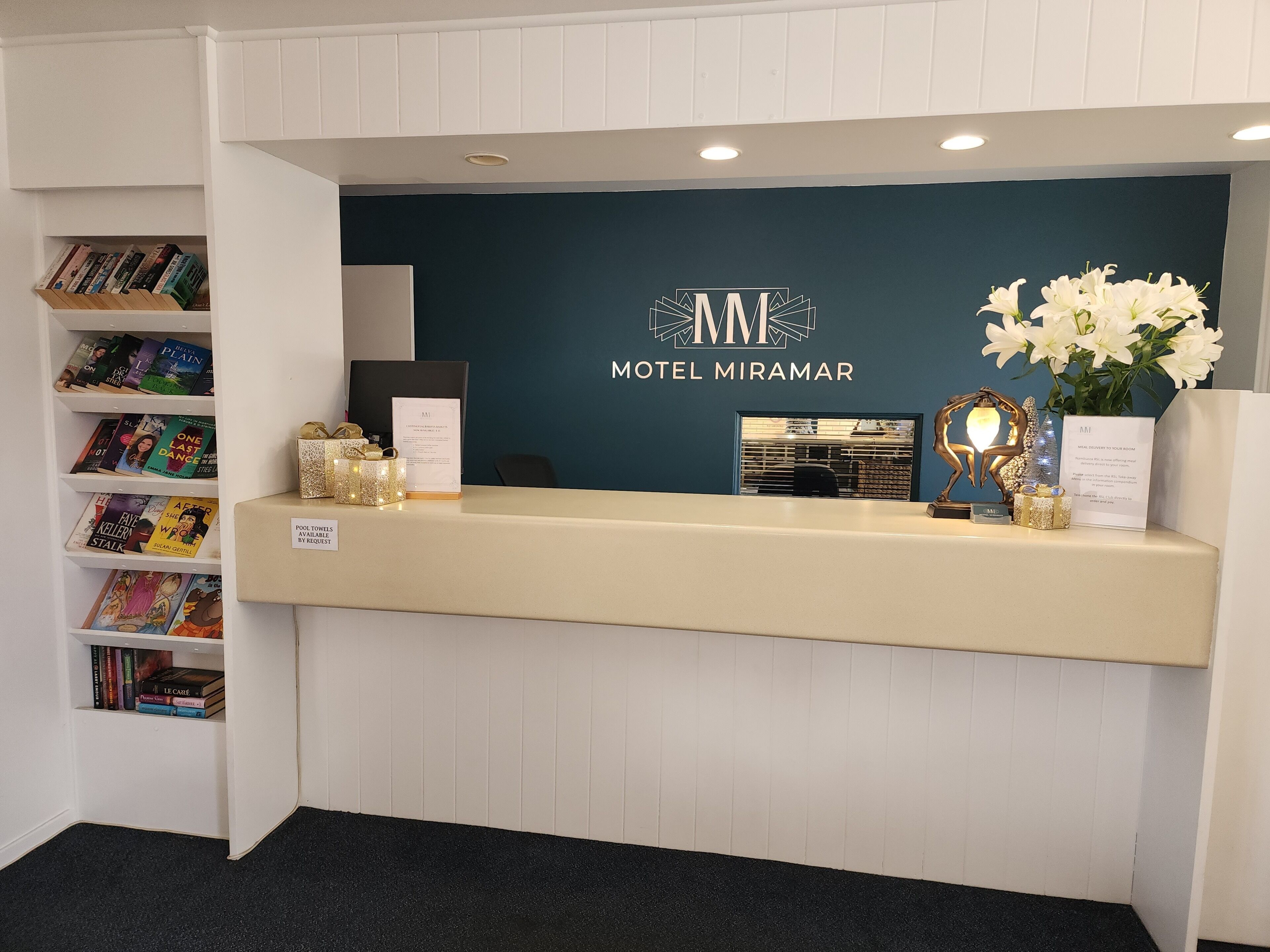 A reception desk at the HOTEL MIRAMAR with a dark blue wall, a white reception area, and a bookshelf with books.