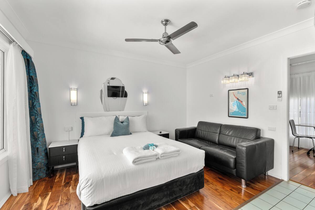 A modern hotel room with a large bed, a black leather sofa, a ceiling fan, and a dining area with a table and chairs.