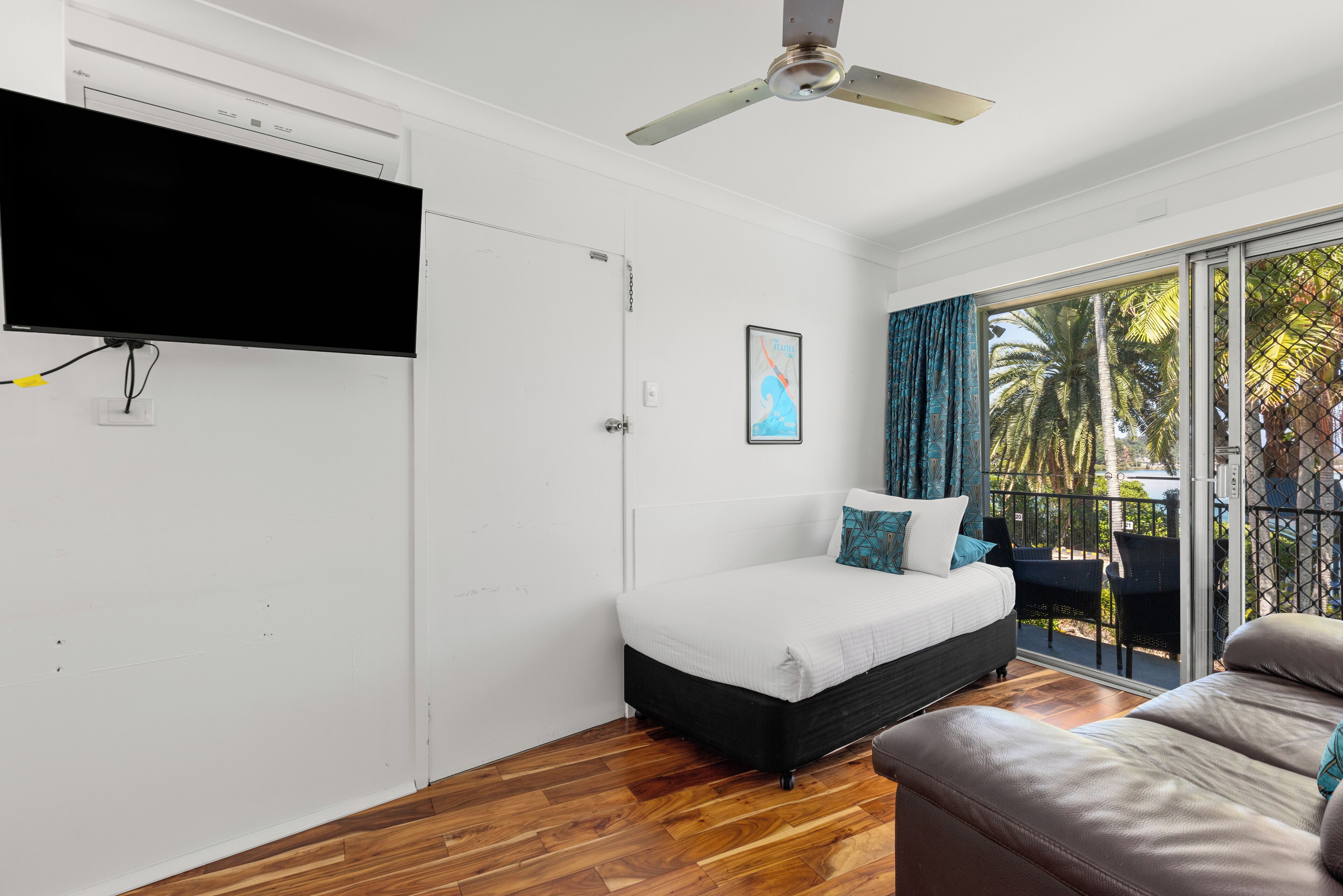 Una habitación con una cama, un sofá, un ventilador de techo, un televisor montado en la pared y un amplio ventanal con vistas a palmeras.
