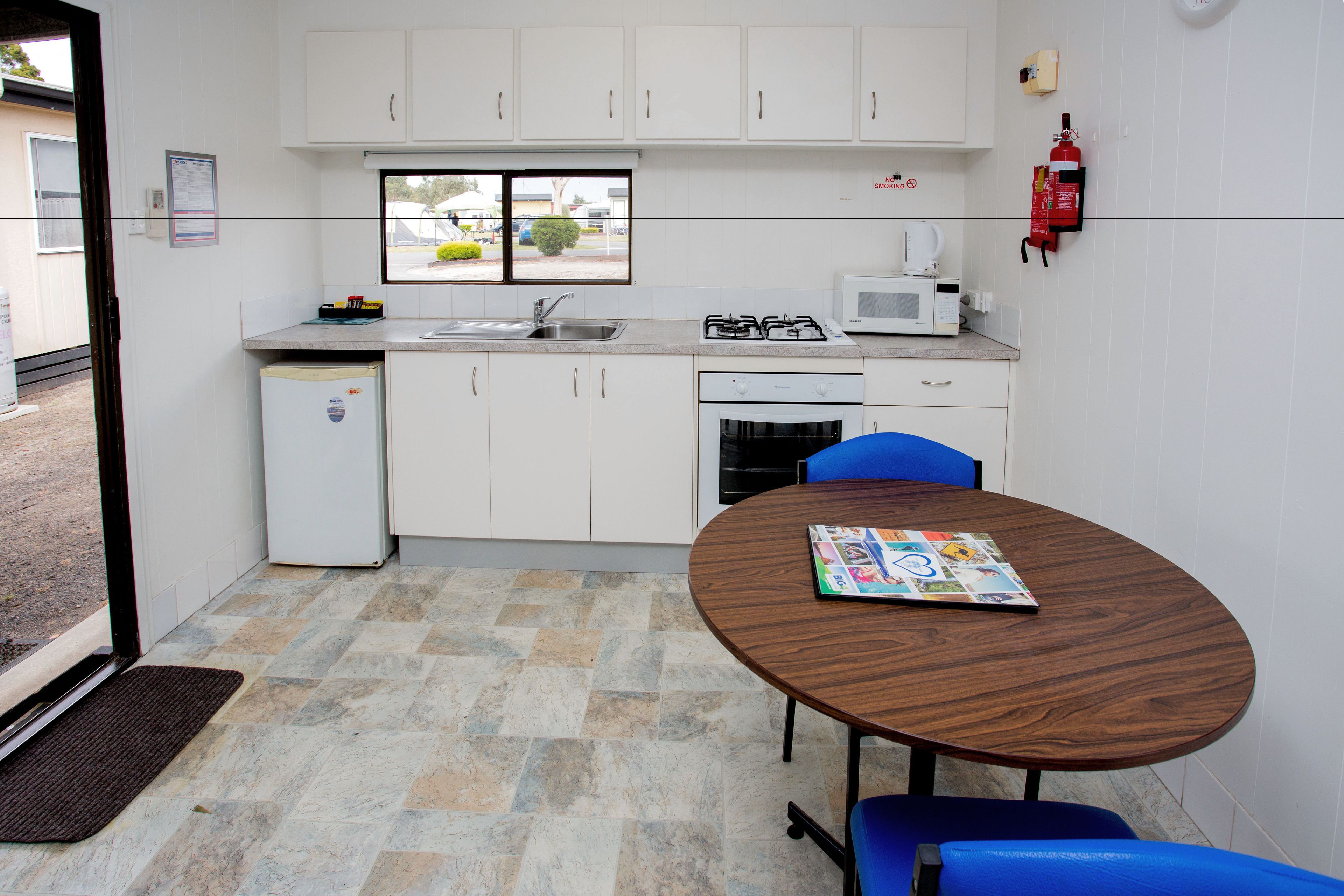 Een compacte keuken met witte kastjes, een magnetron, een gasfornuis en een spoelbak. Er is een kleine koelkast, een tafeltje met een tijdschrift, en blauwe stoelen.