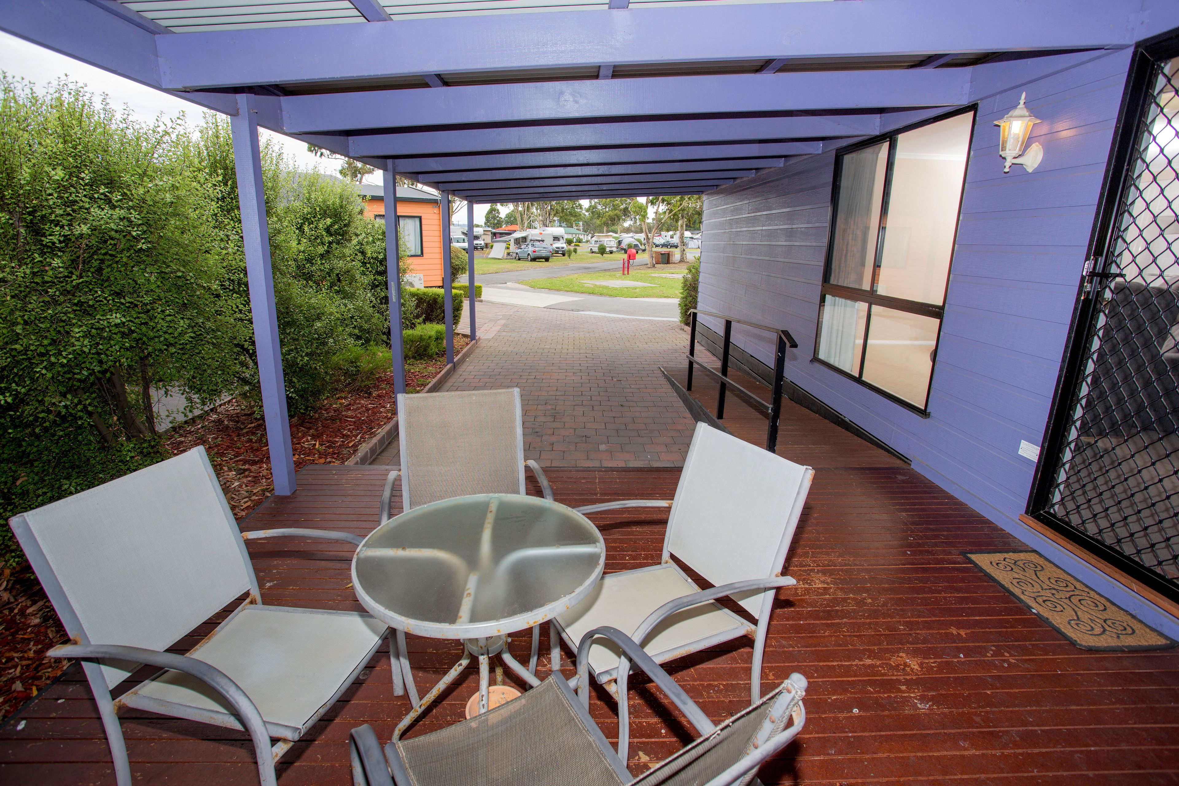A covered outdoor seating area with a table and four chairs, a pathway leading to a building, and a view of parked vehicles and greenery.
