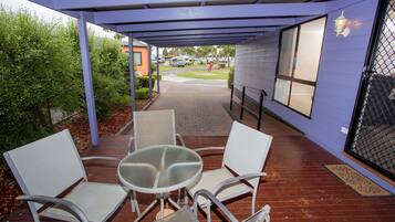 A covered outdoor seating area with a table and four chairs, a pathway leading to a building, and a view of parked vehicles and greenery.
