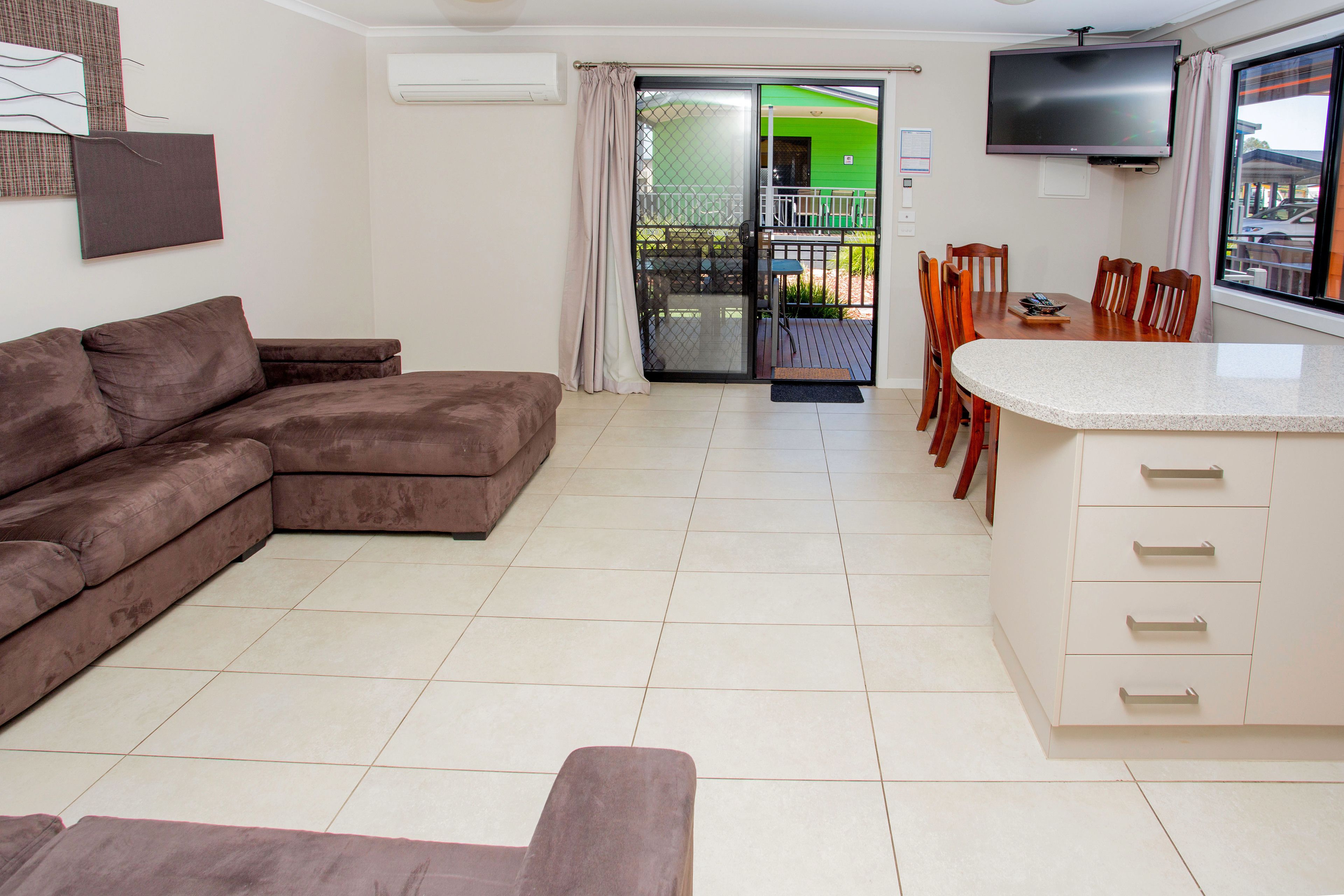 A living room with a brown sectional sofa, a dining area with wooden chairs and a table, and a kitchen area with a white countertop and cabinets.
