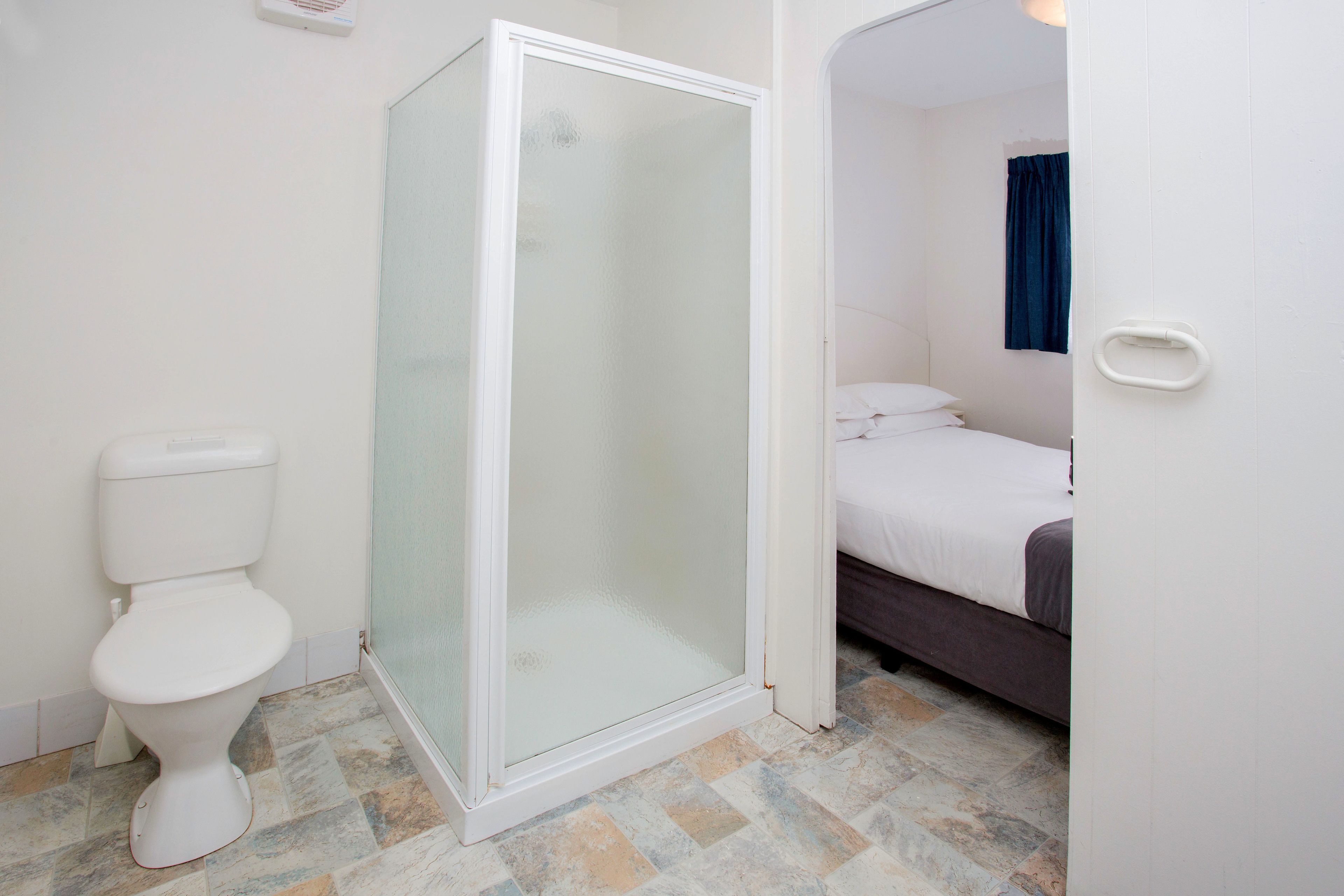 A bathroom with a glass shower enclosure, a toilet, and a bed with a blue curtain.