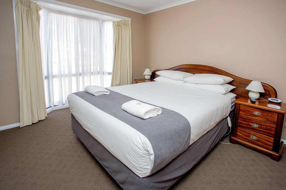 Een net opgemaakt bed met een grijs-witte dekbedovertrek, een houten nachtkastje met een lamp en een raam met dunne gordijnen.