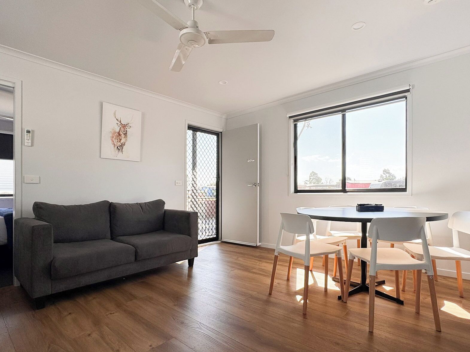 A modern living room with a grey sofa, a dining table with chairs, a ceiling fan, and a large window.