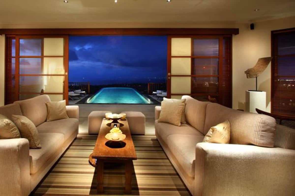 A modern living room with two sofas facing a pool, a coffee table, and large windows with a view of the pool at night.