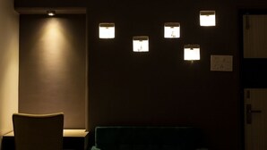 A dimly lit room with a single chair and a couch. There are five wall-mounted light fixtures, one of which is illuminated. A door is visible on the right side. - Simi International - The Imperial (Bidhannagar)
