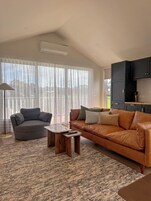 Sala de estar com sofá, mesa de centro e luminária. Há uma janela ampla com cortinas leves.