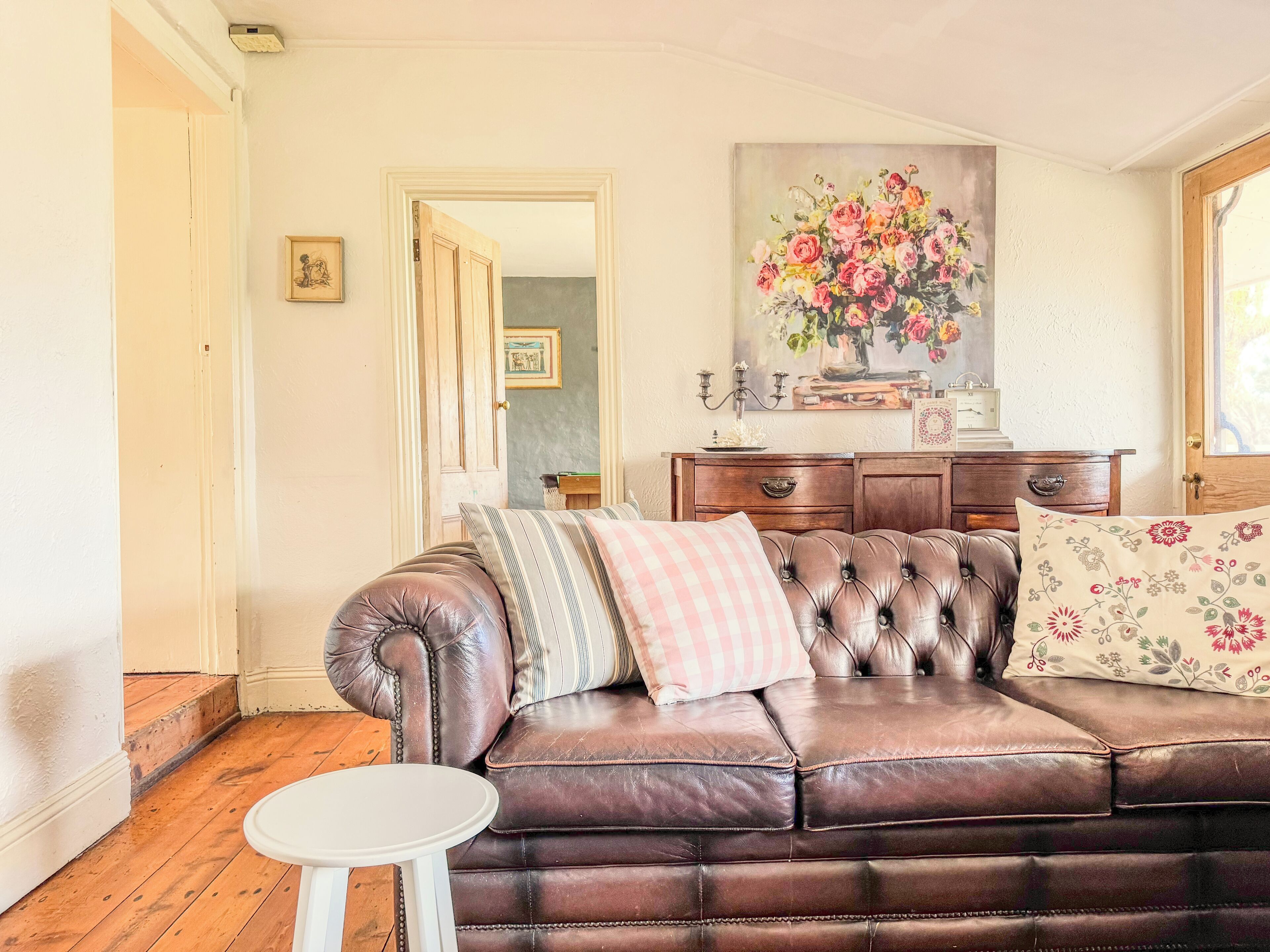 Ein Wohnzimmer mit einem braunen Ledersofa, einem kleinen weißen Tisch und einer Holzkommode mit einem floralen Gemälde darüber.