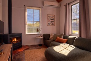 A cozy living room with a wood-burning stove, a sofa, a coffee table, and a window with blinds. - Clonmara Country House & Cottages (Port Fairy)