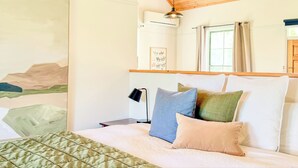 A bedroom with a bed, pillows, a nightstand, a lamp, a window with curtains, a wall-mounted air conditioner, and a framed picture. - Clonmara Country House & Cottages (Port Fairy)