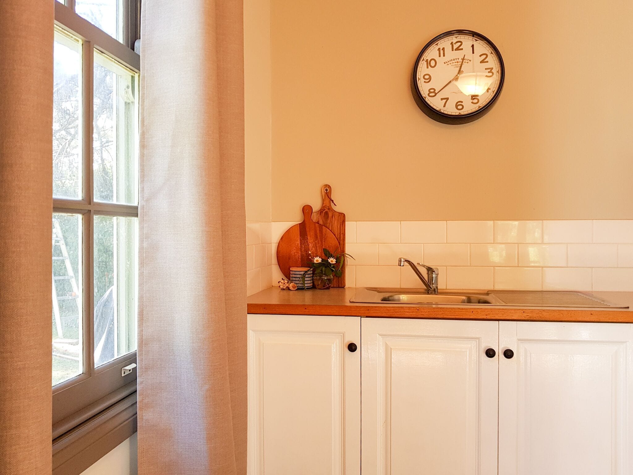 Eine Küche mit weißen Schränken, einer Holzausziehtafel, einem Spülbecken, einer Uhr und einem Fenster mit Vorhängen.