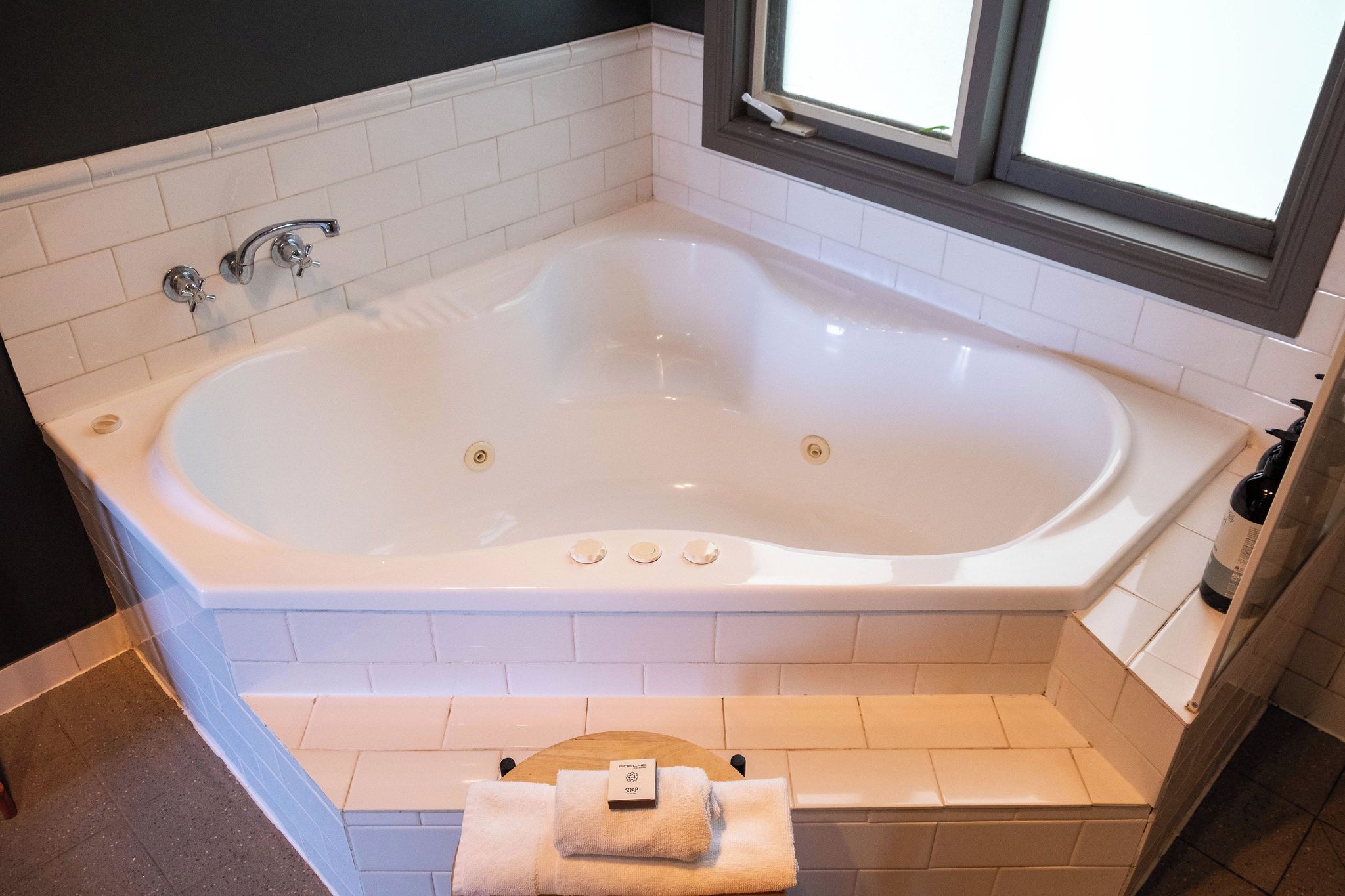 A large freestanding bathtub with a window above it, a towel on the floor, and a soap dispenser on the wall.