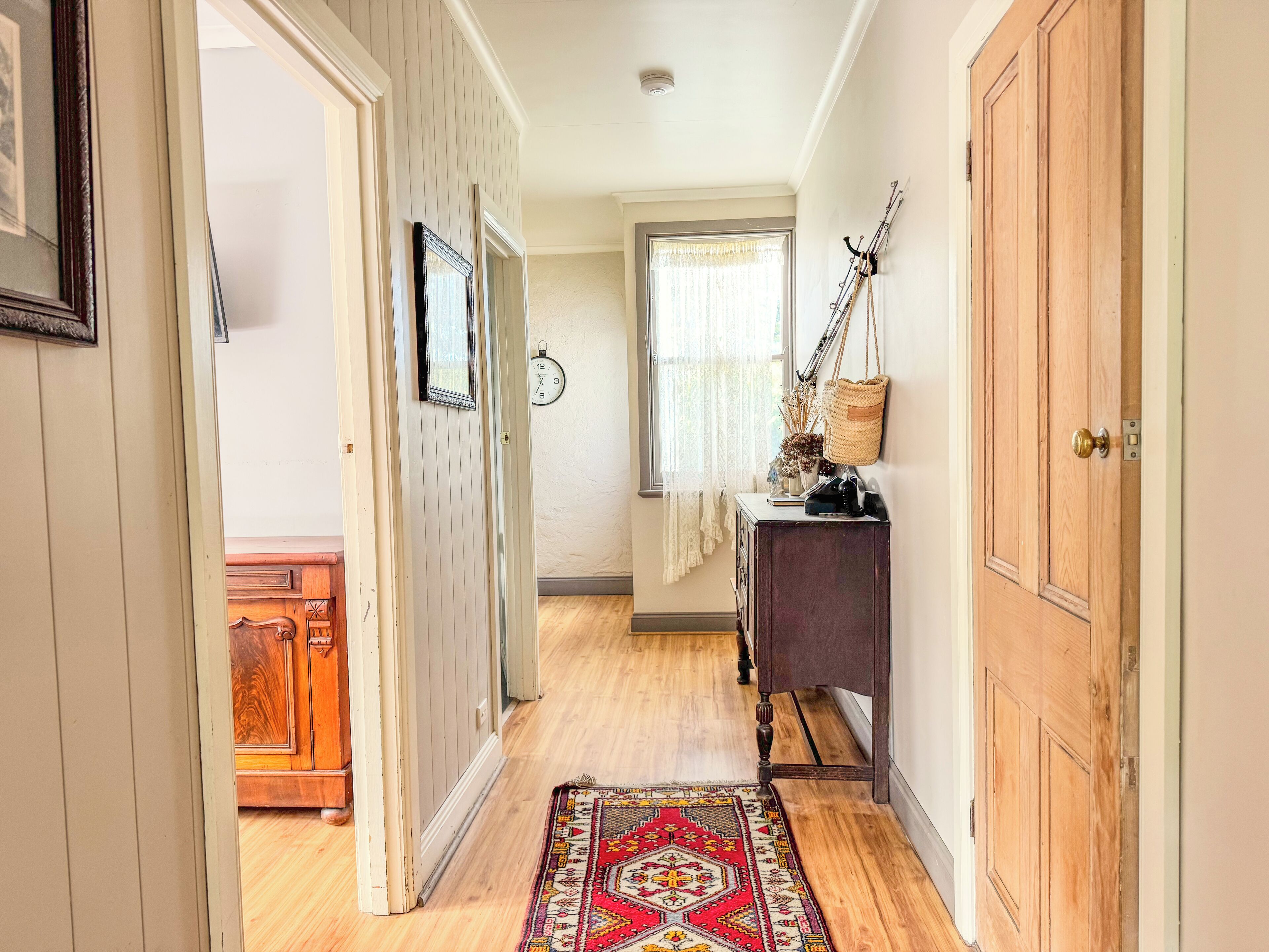 Un couloir avec un plancher en bois, un tapis à motifs, une armoire en bois, une horloge et une porte dotée d’une poignée en laiton.