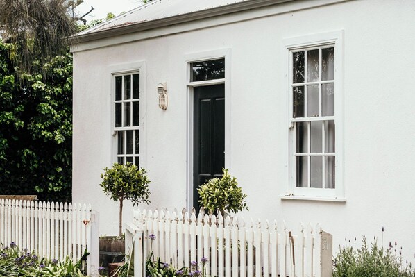 A white building with two windows and a door, a white picket fence, and small trees in front.