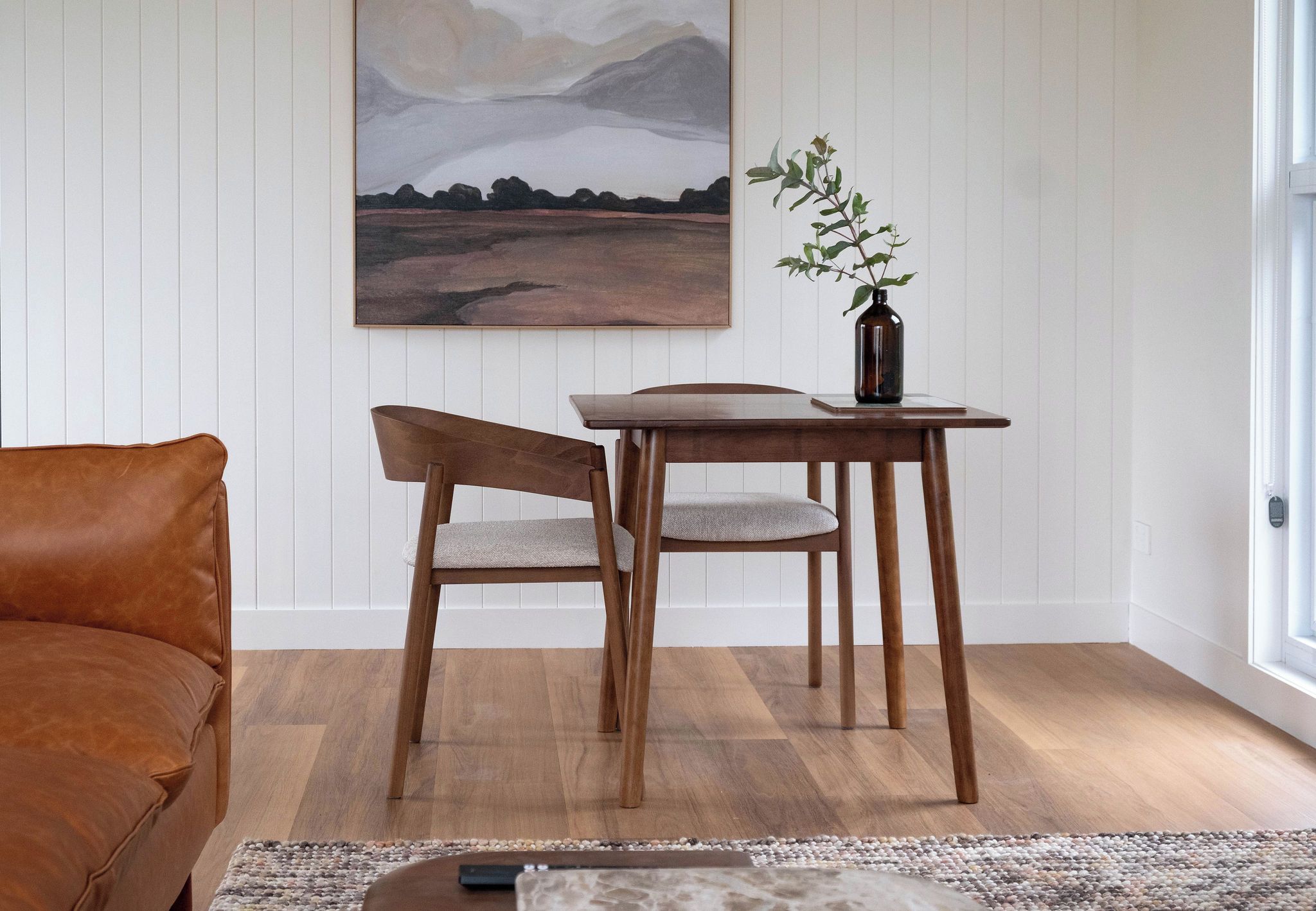 A room with a wooden dining table, two chairs, a brown leather sofa, a painting on the wall, and a vase with a plant on the table.