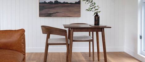 A room with a wooden dining table, two chairs, a brown leather sofa, a painting on the wall, and a vase with a plant on the table.