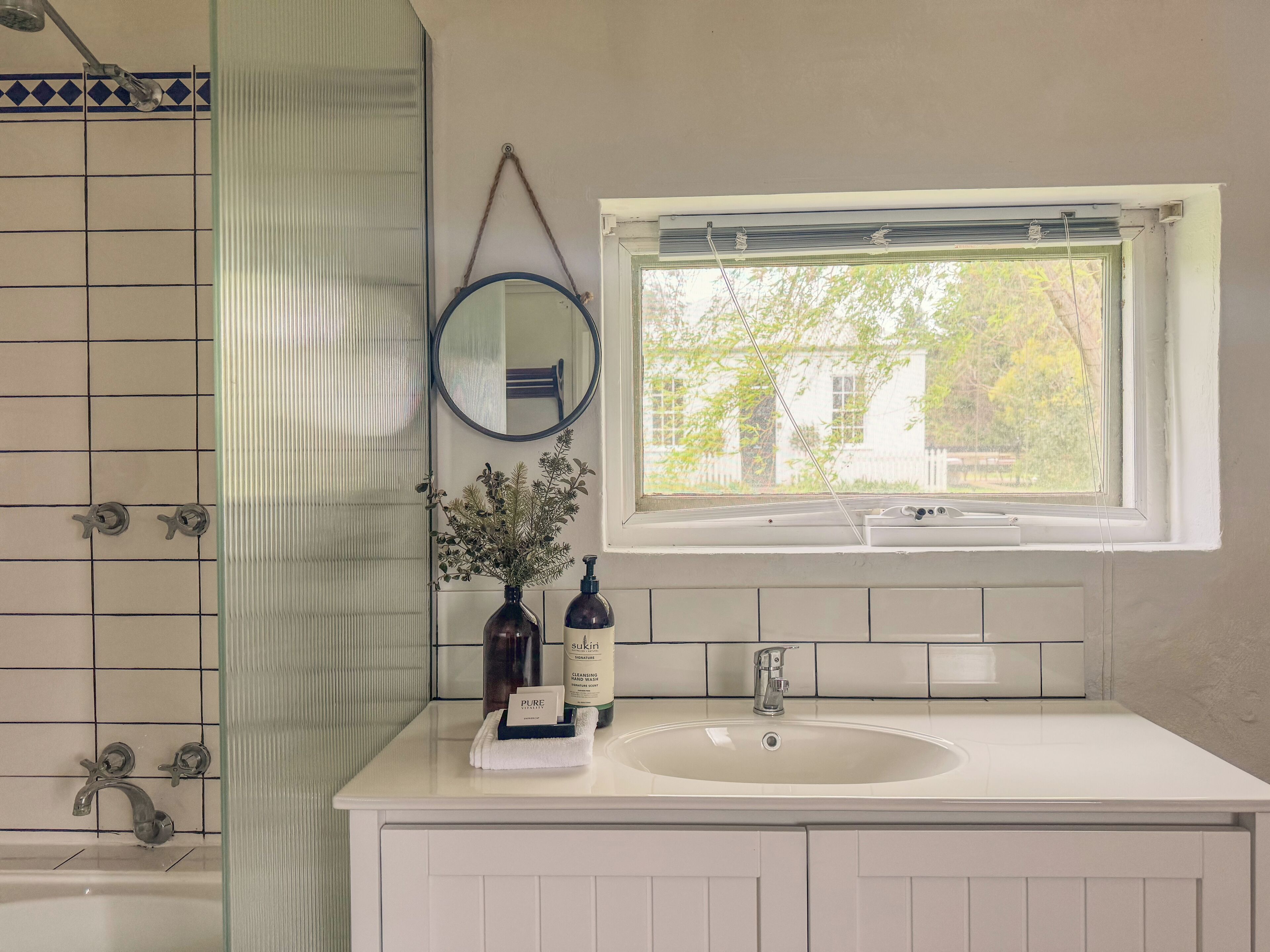 A bathroom with a white sink, a mirror, a window with a view of greenery, and a shower area with a tiled wall.