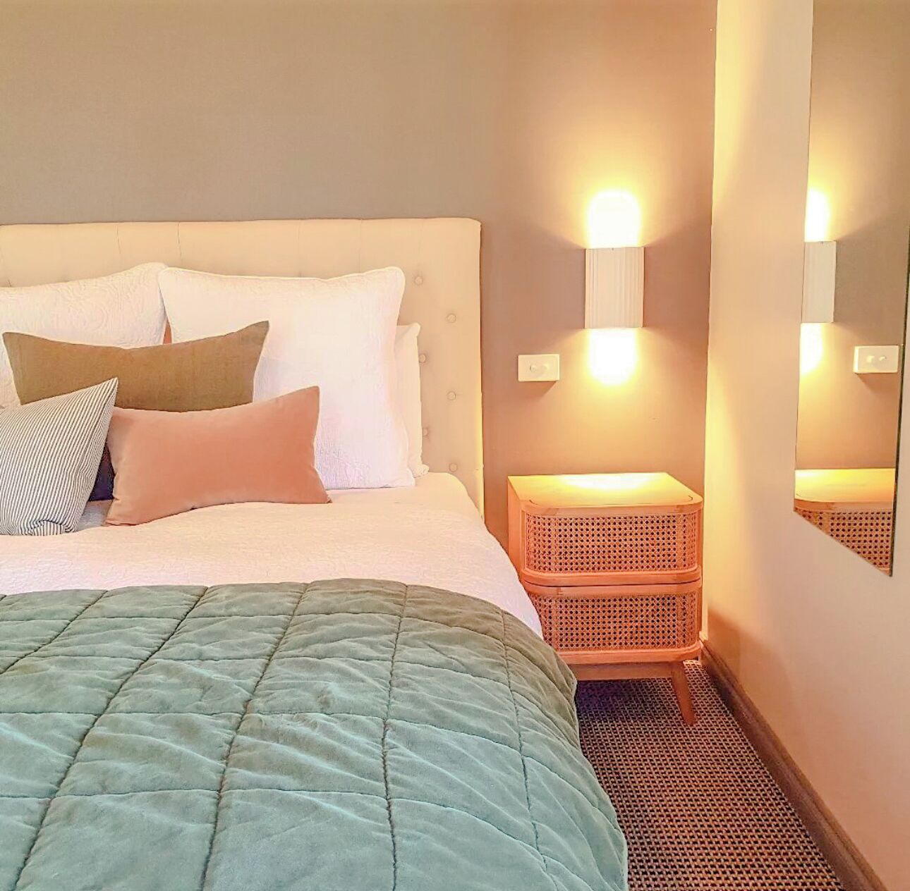 A neatly made bed with a green quilt, multiple pillows, and a bedside table with a woven basket.