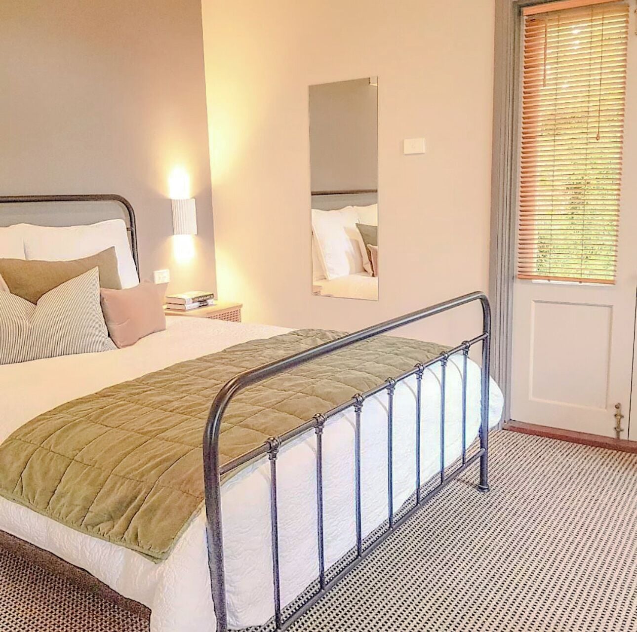 A neatly made bed with a metal headboard, a bedside lamp, a mirror on the wall, and a window with blinds.