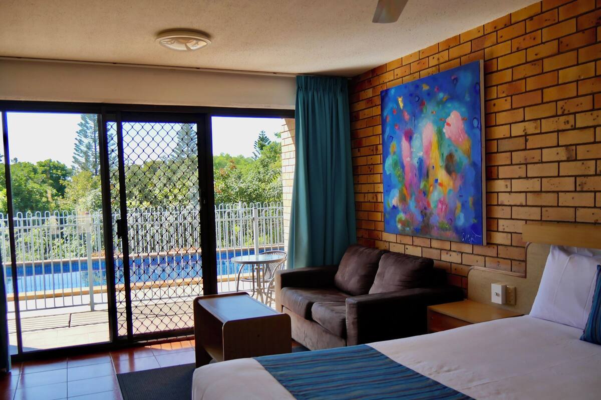 Une chambre d’hôtel avec un lit, un canapé, une petite table et une vue sur une piscine, visible à travers une porte vitrée.