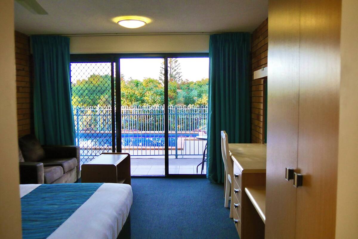 Une chambre d’hôtel équipée d’un lit, d’un bureau, d’une chaise et offrant une vue sur une piscine, visible à travers une porte vitrée.