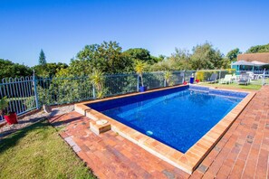 Une piscine rectangulaire dont le bord est recouvert de carreaux, entourée d’une terrasse en briques et d’une clôture en métal.