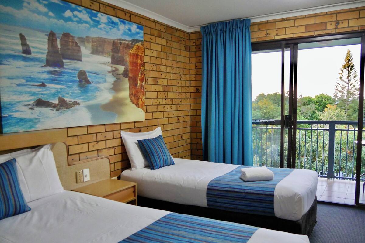 Une chambre d’hôtel avec deux lits, un grand tableau représentant un paysage côtier et un balcon donnant sur un espace verdoyant.