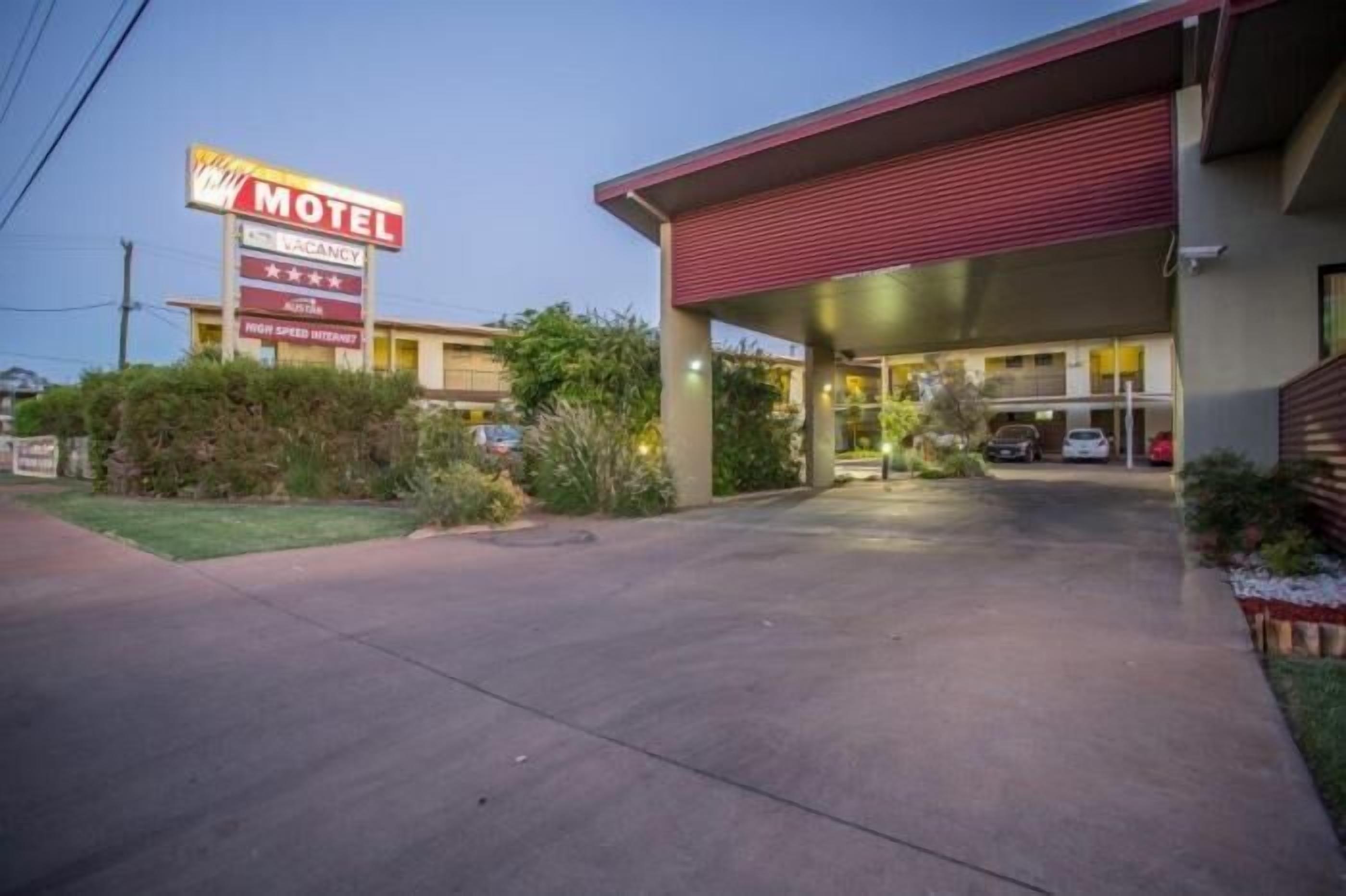 Um motel com toldo vermelho, placa indicando quartos disponíveis e estacionamento com carros.
