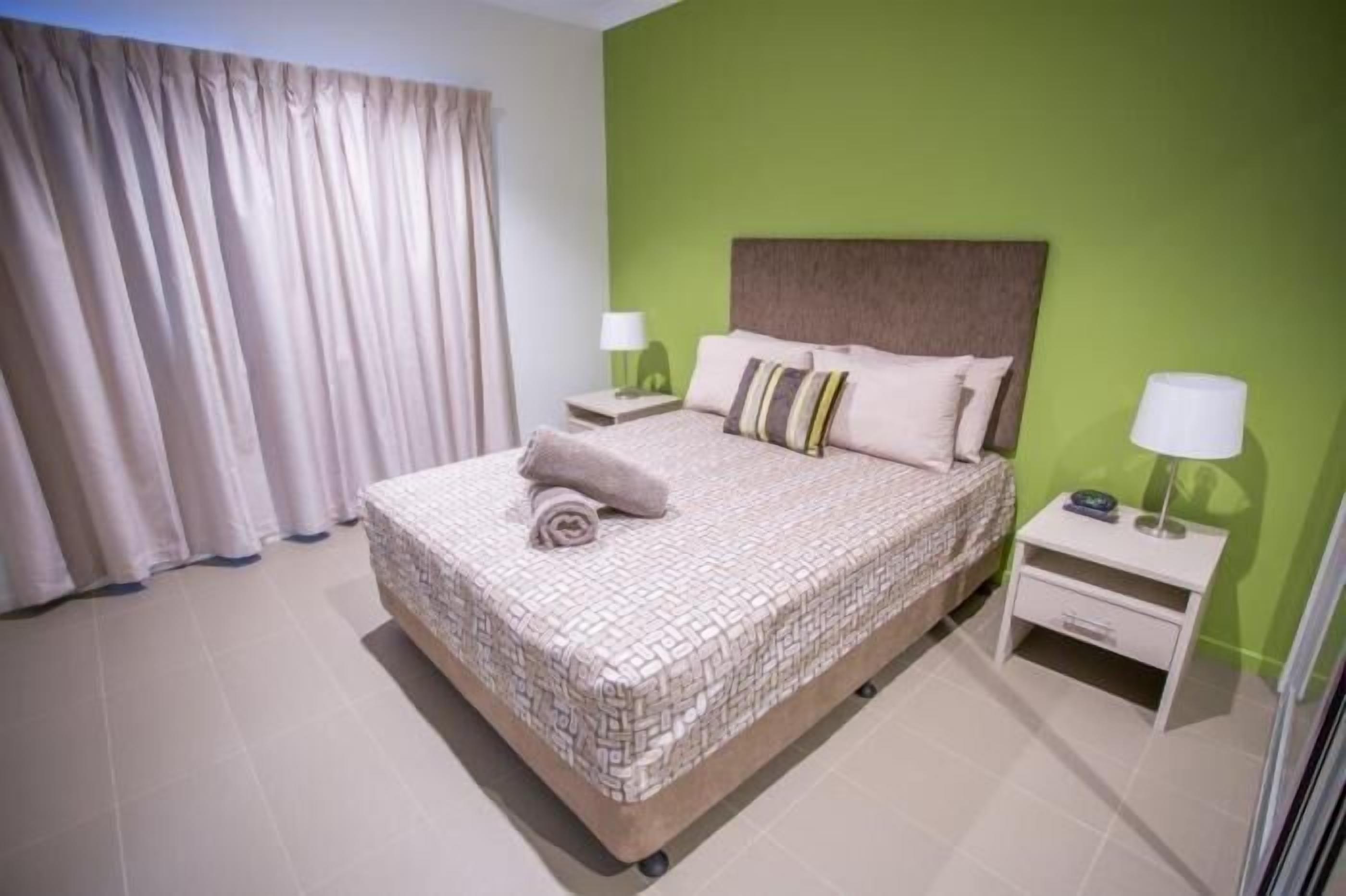 A neatly made bed with a patterned bedspread, two bedside tables with lamps, and a green accent wall.