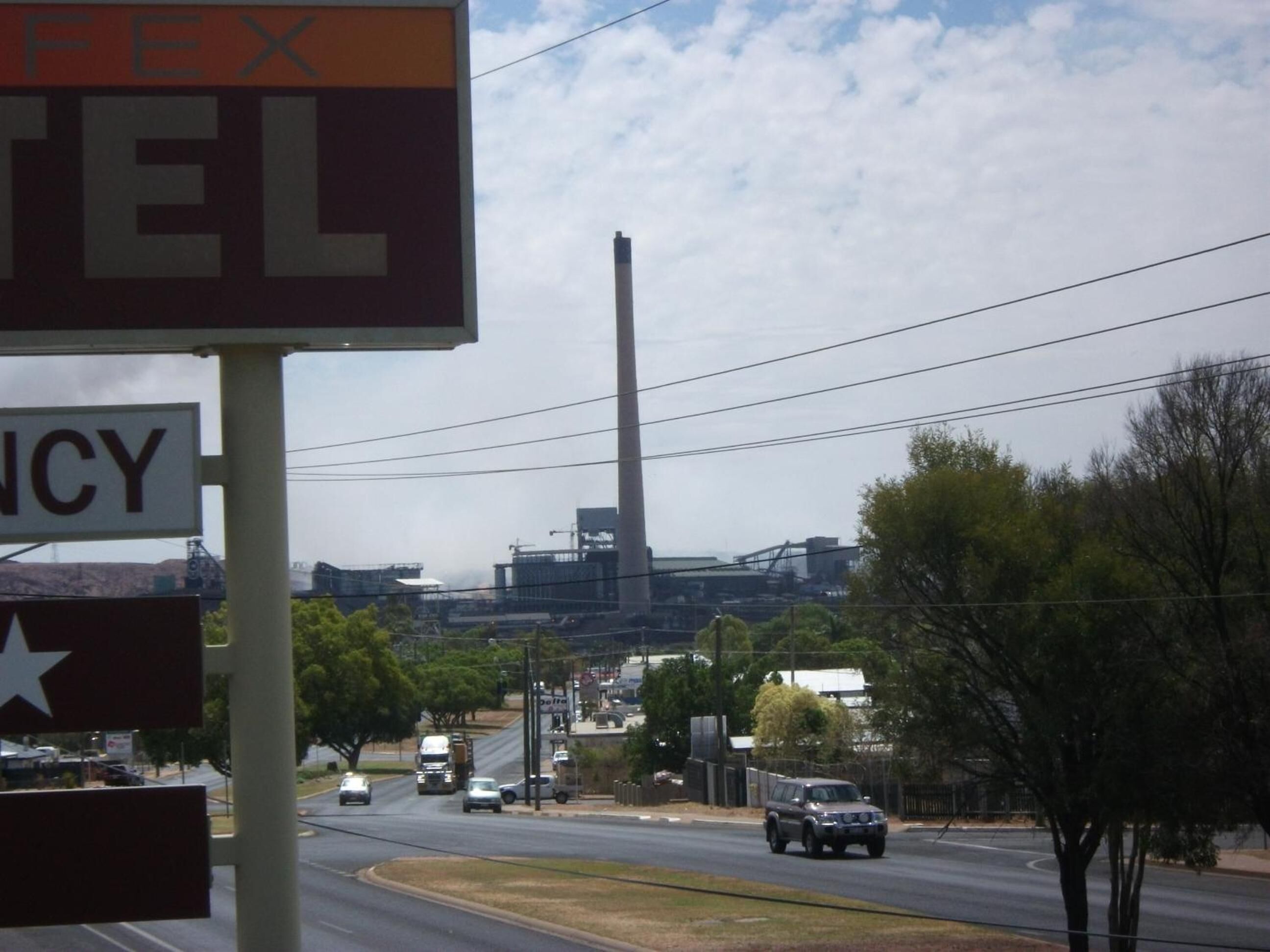 A road with vehicles, a large industrial building in the background, and a sign with partially visible text.