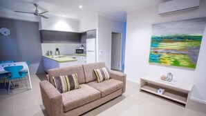 A modern living room with a beige sofa, a dining table with blue chairs, a kitchen area, and a painting on the wall. - Spinifex Motel & Serviced Apartments (Townview)