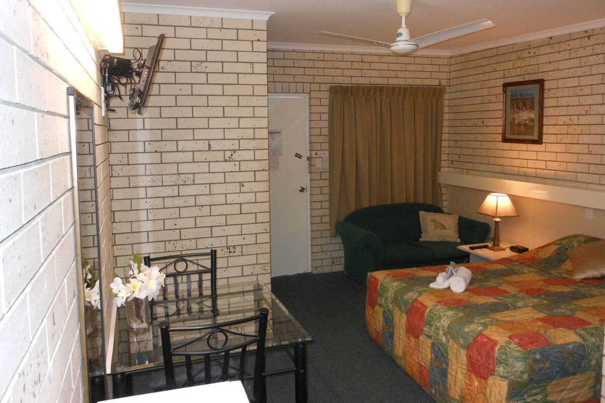 A room with a brick wall, a bed with a colorful quilt, a green armchair, a table with chairs, a ceiling fan, and a lamp.