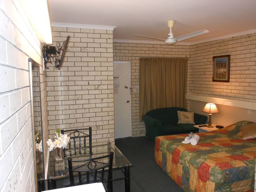 A room with a brick wall, a bed with a colorful quilt, a green armchair, a table with chairs, a ceiling fan, and a lamp.