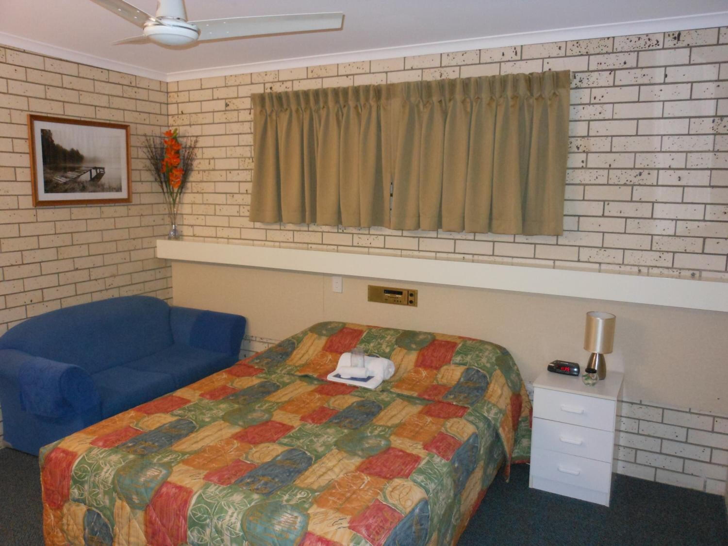 A room with a bed, a blue sofa, a window with curtains, a ceiling fan, and a wall-mounted radio.