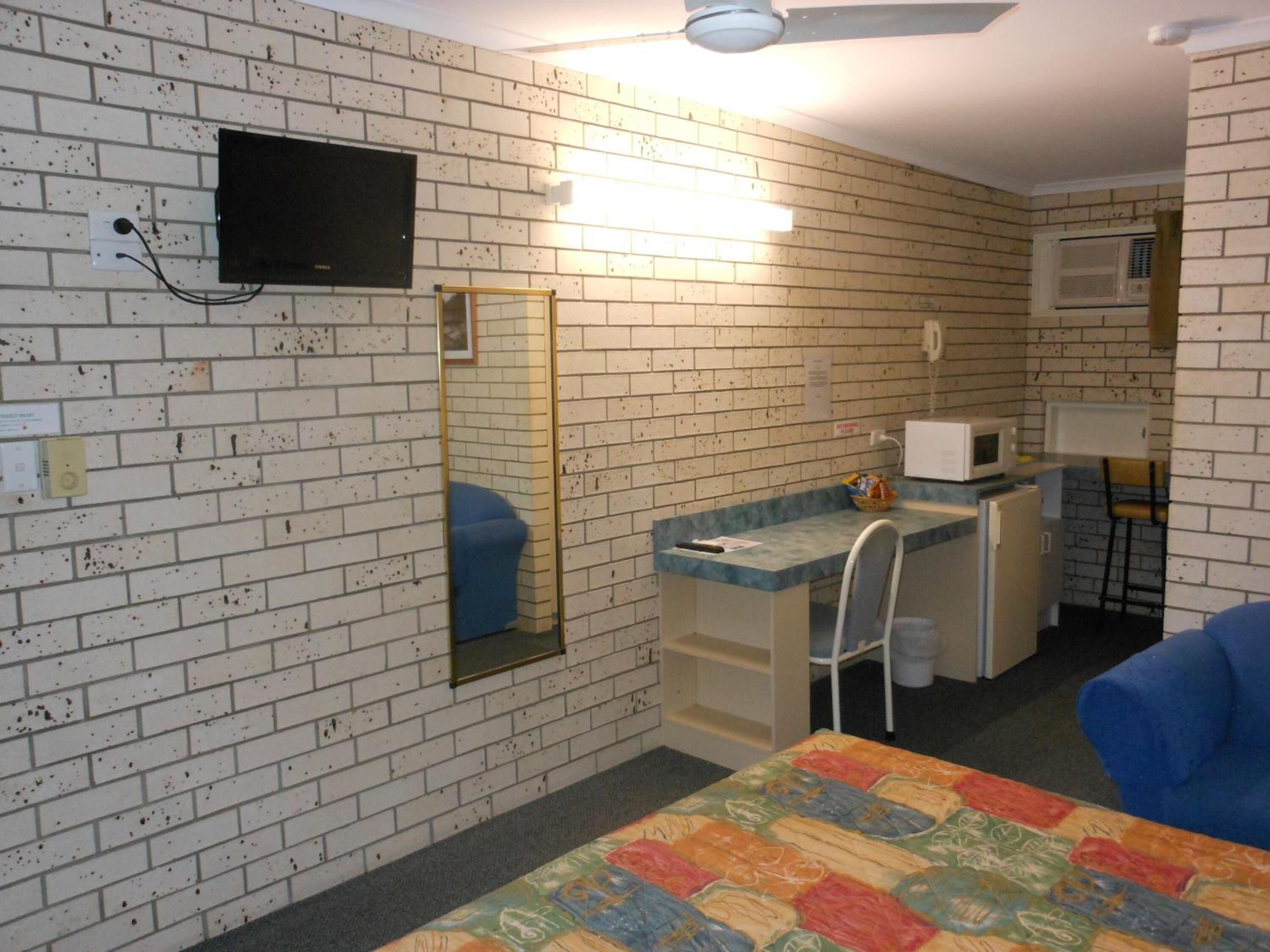 Een kamer met een wand in baksteenmotief, een bed met een kleurrijke quilt, een bureau met een stoel, een magnetron en een aan de muur bevestigde televisie.