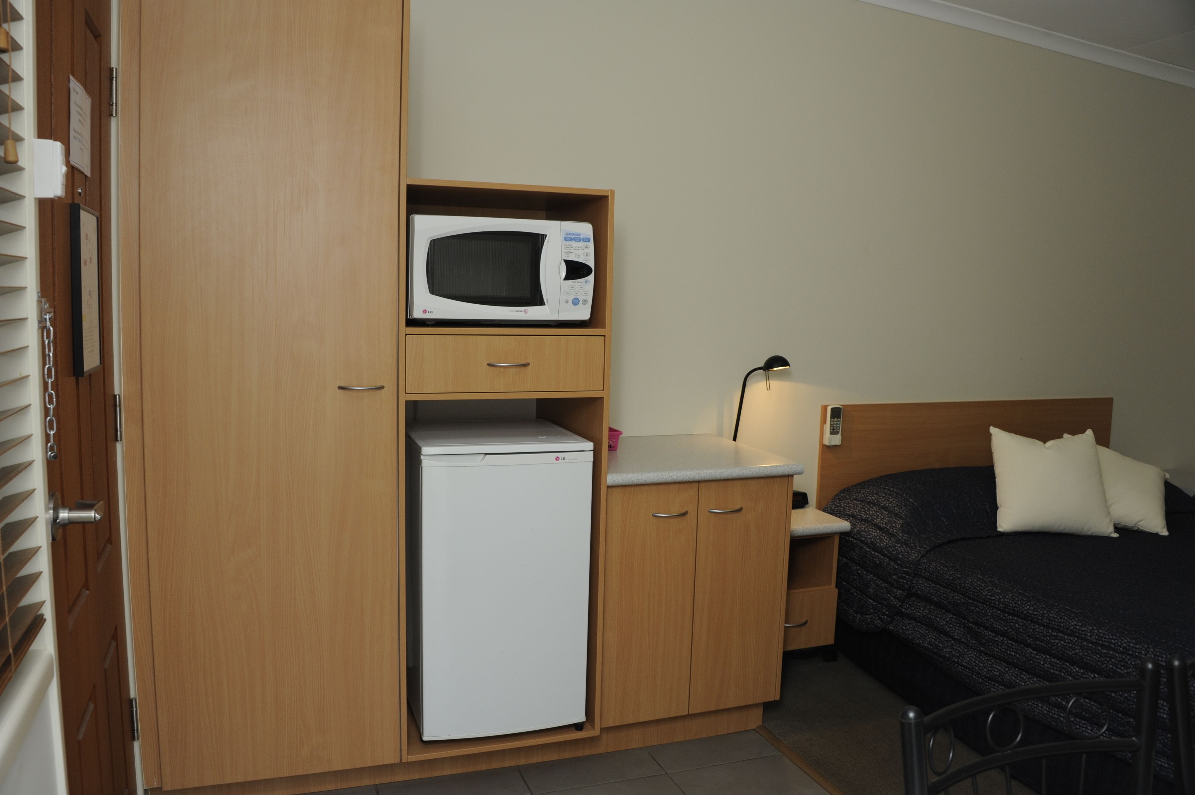 A compact room with a bed, a bedside table with a lamp, a microwave, and a mini-fridge.